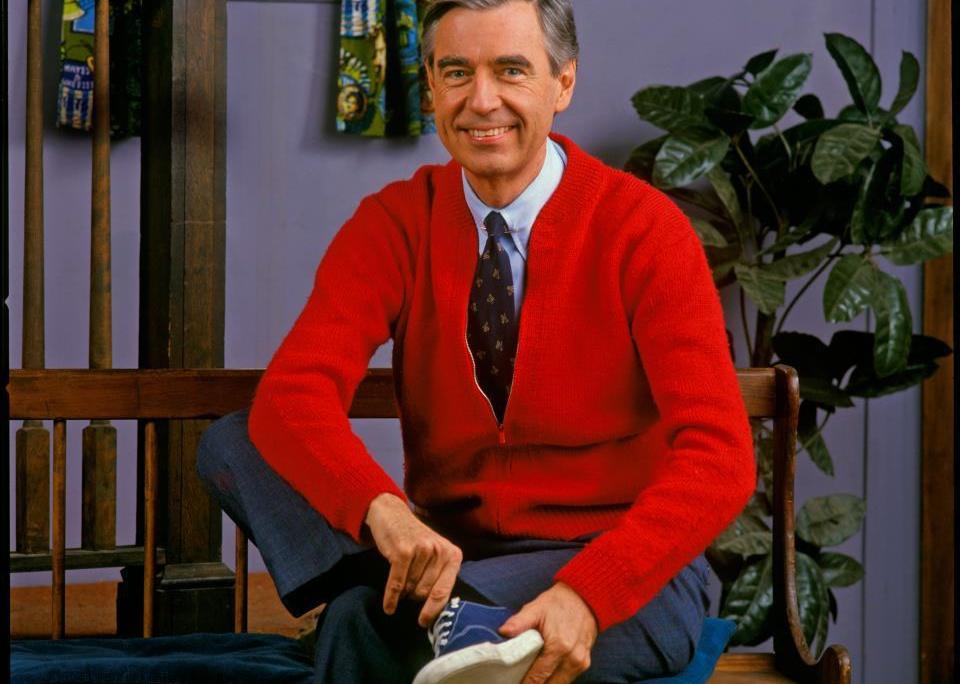 Fred Rogers putting on his shoe in a red sweater.
