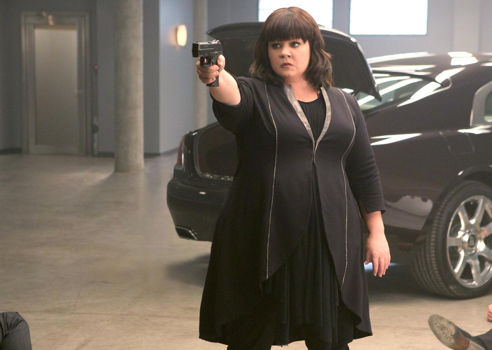 Melissa McCarthy in all black pointing a gun in a parking garage.