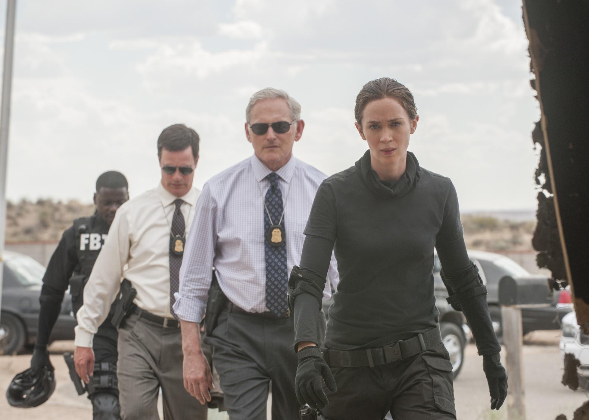 Emily Blunt and Victor Garber walk in a line of men in all black.