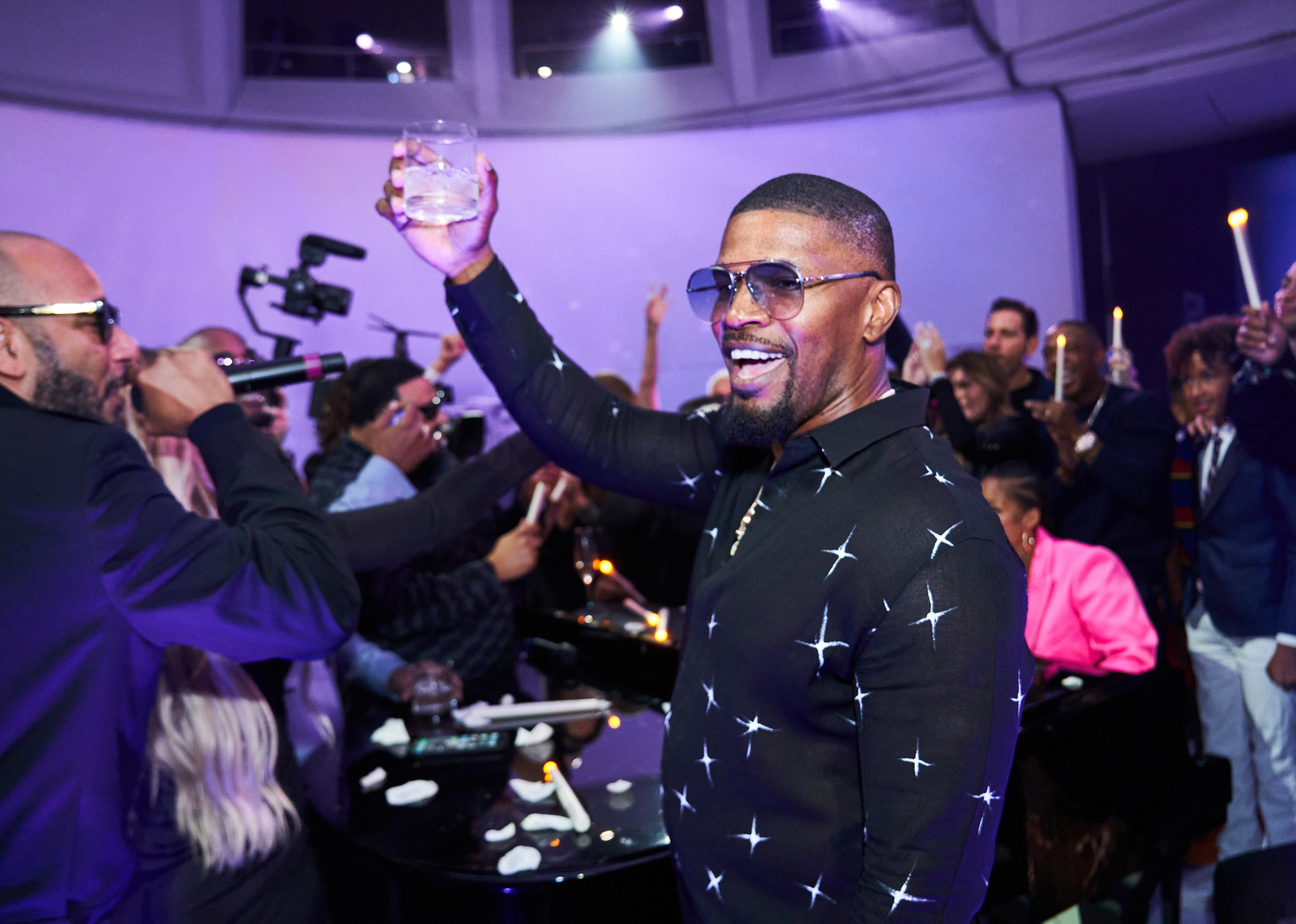 Jamie Foxx in a black shirt and sunglasses holding up a drink. 