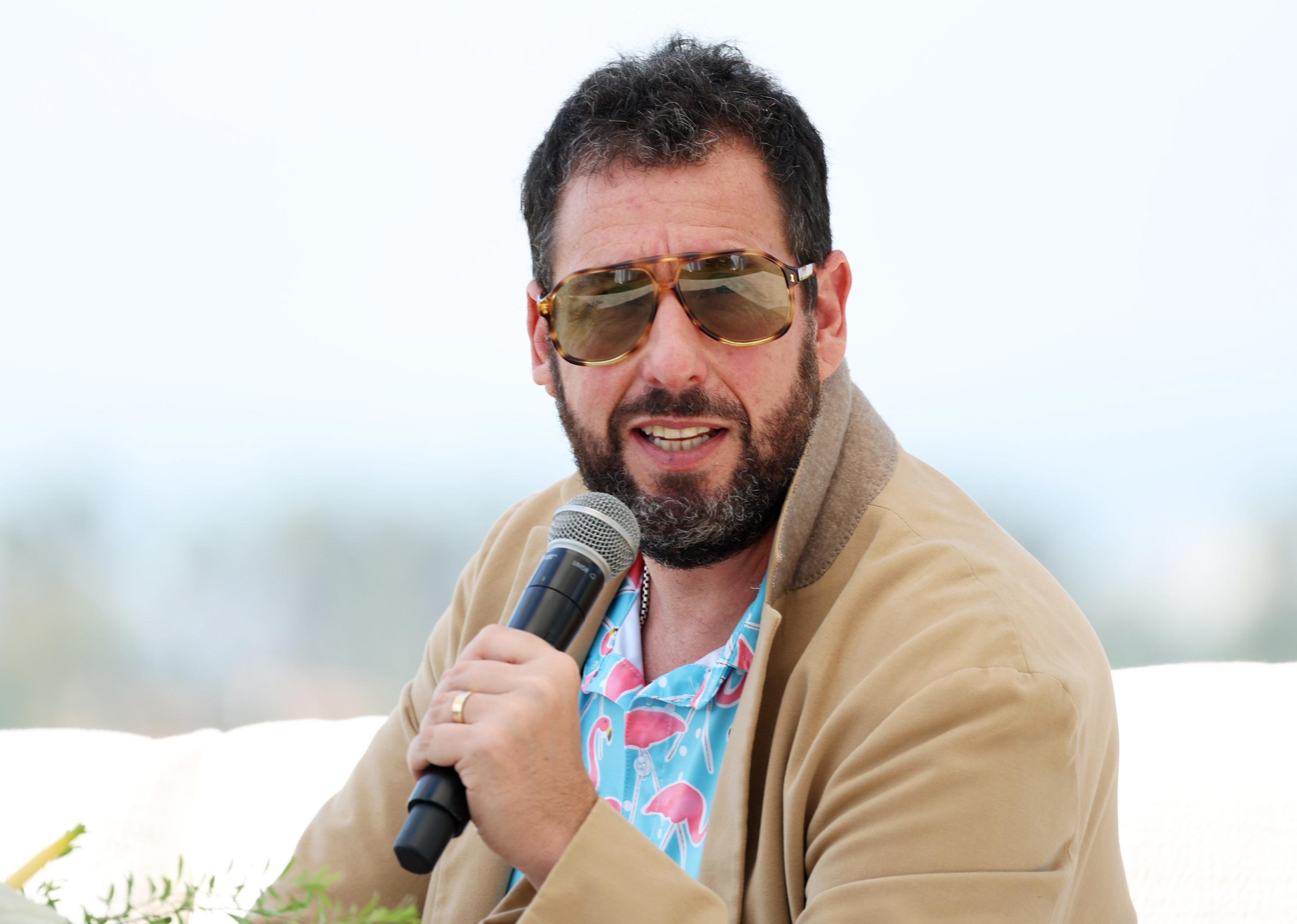 Adam Sandler in a flamingo shirt and sunglasses with a microphone.