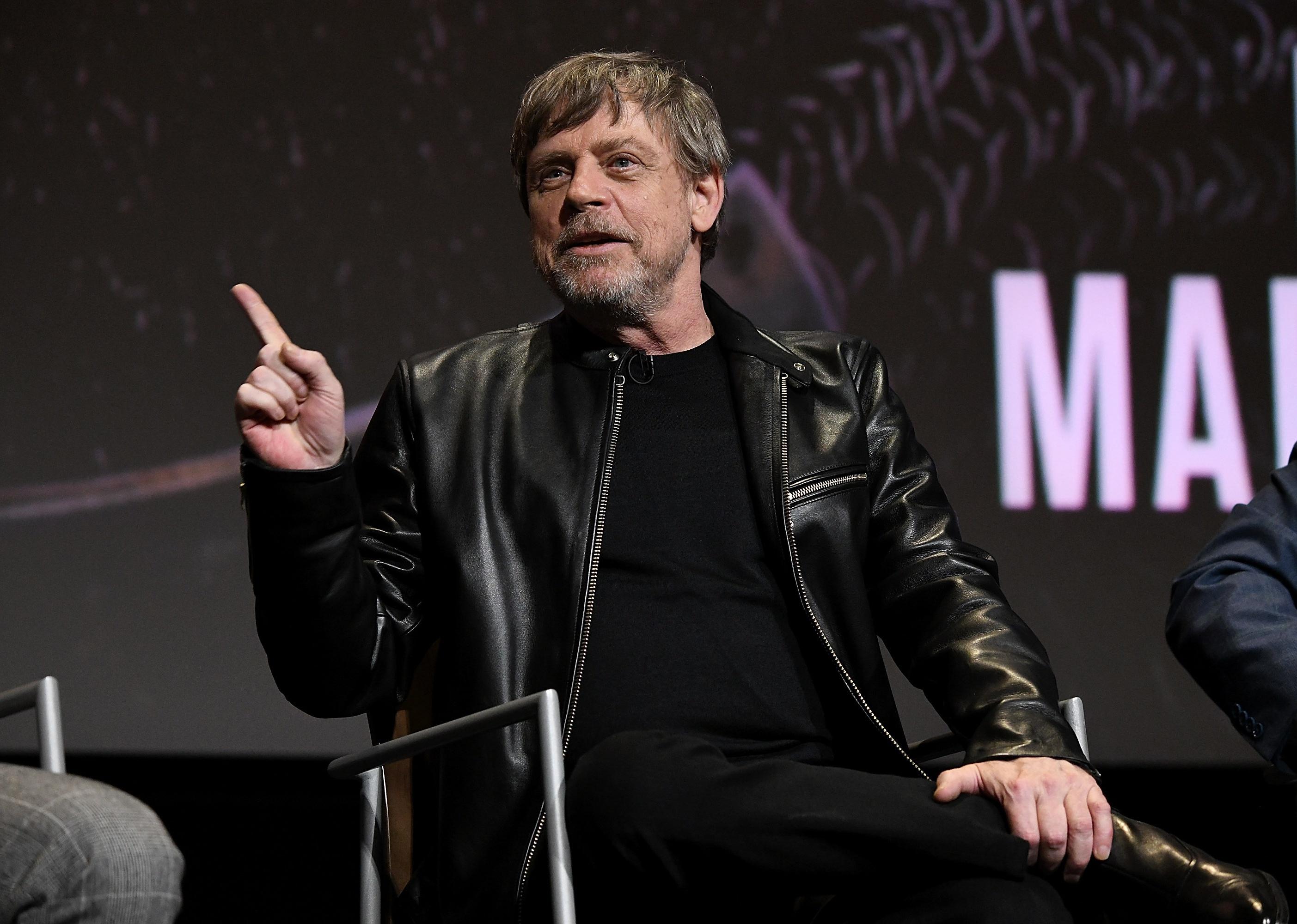 Mark Hamill in a black leather jacket sitting onstage.