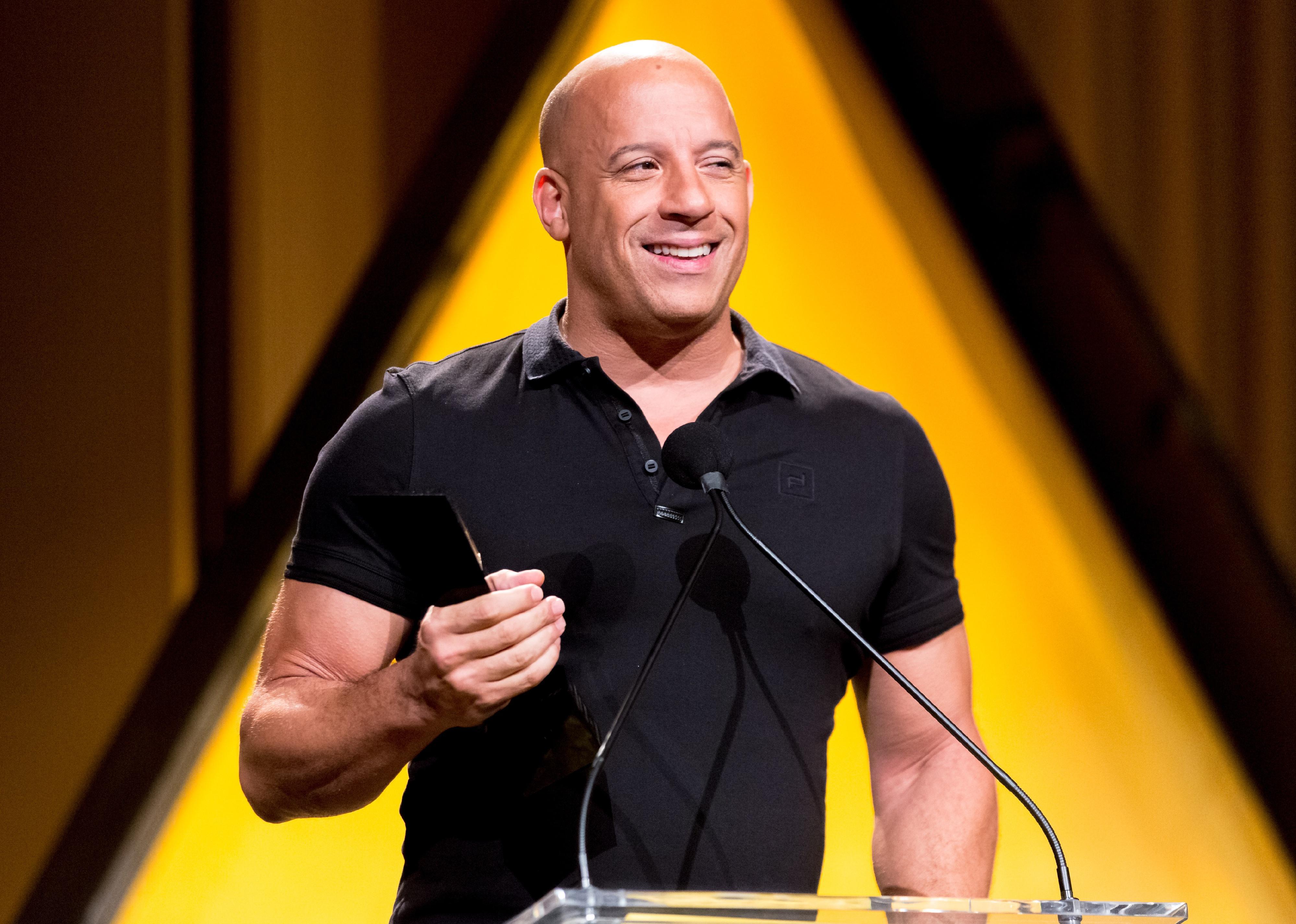 Vin Diesel in a black polo shirt behind a podium.