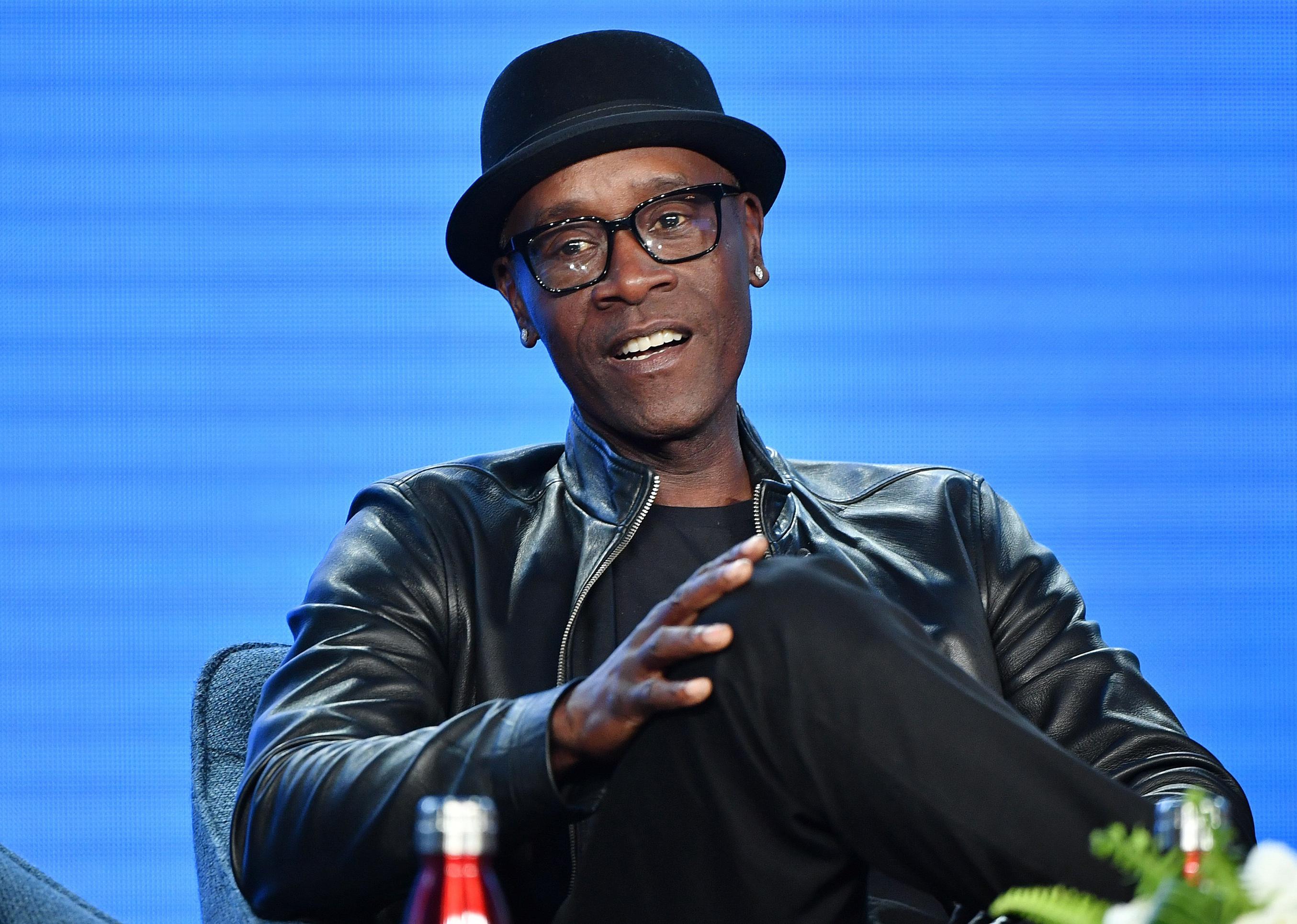 Don Cheadle sitting onstage in a black leather jacket and hat.