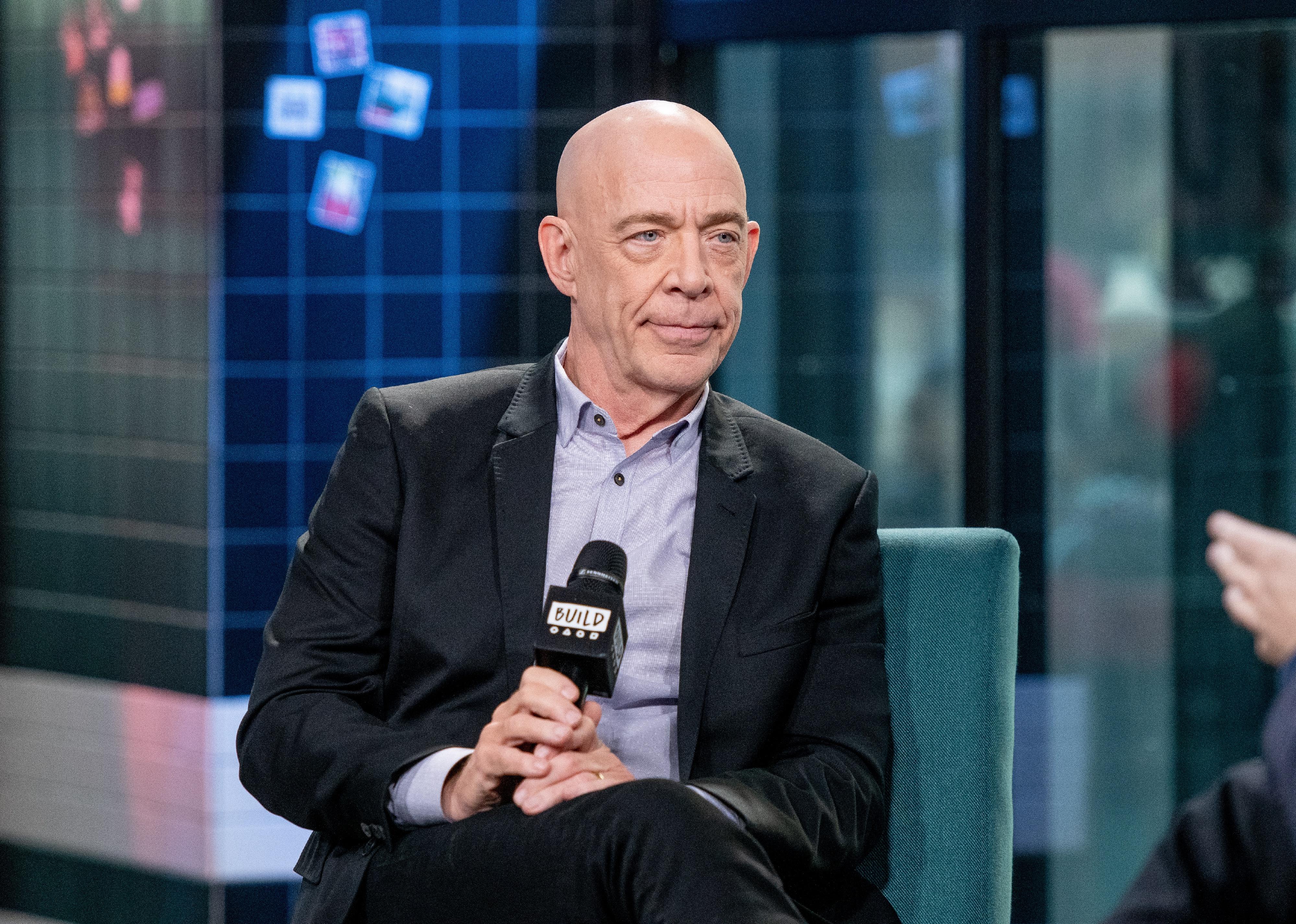 J.K. Simmons in a black jacket holding a microphone onstage.