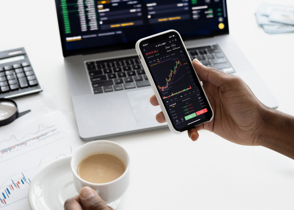 A person's hands holding a cup of coffee and a phone with crypto and bitcoin up on phone and computer.