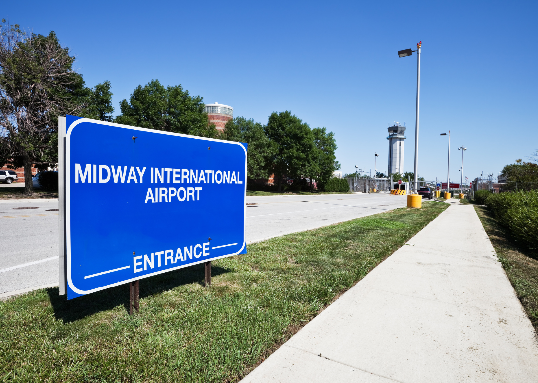A blue sign designating the entrance the the Midway International Airport with the control tower in the background.