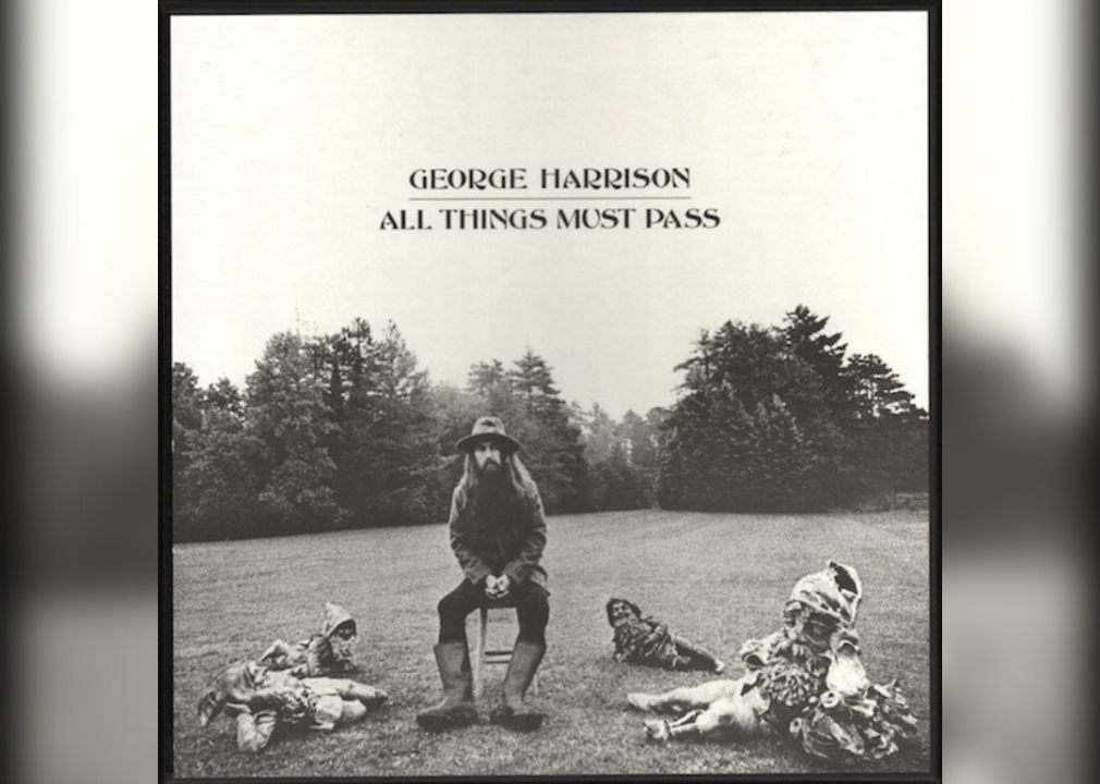 George Harrison sits in a chair in a field surrounded by gnome-like figures lying on the ground.