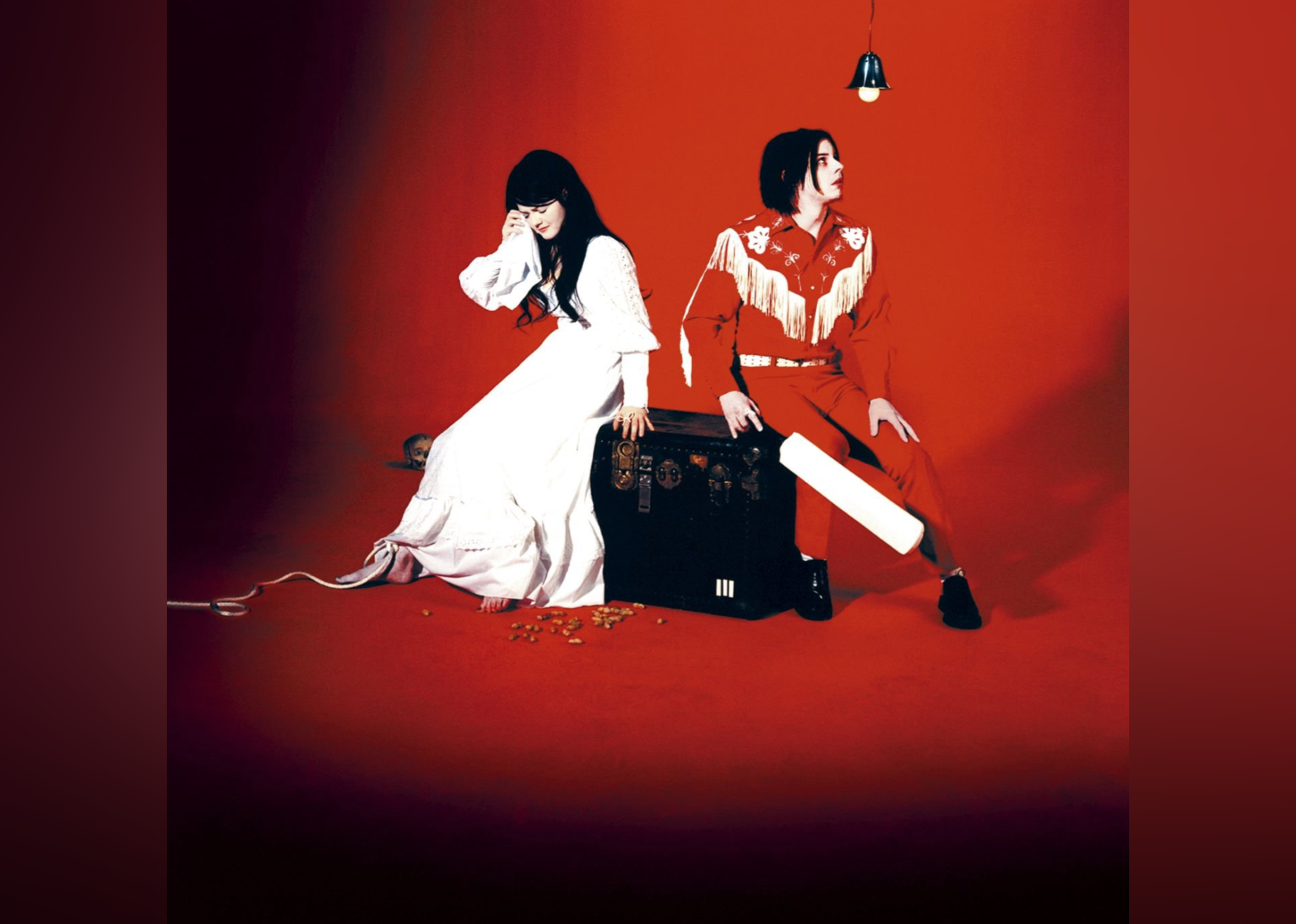 Meg White wearing a white dress and crying sitting on a trunk next to Jack White in a red western outfit holding a paddle.