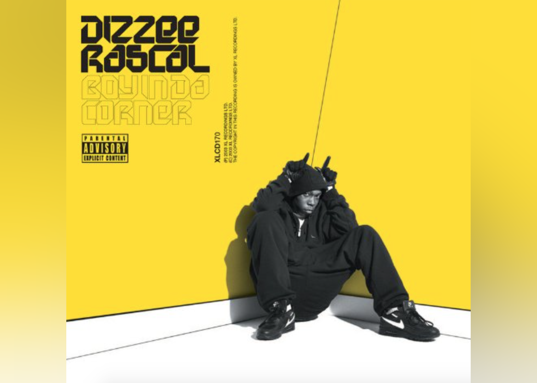 Dizzee Rascal sitting in the corner of a yellow room wearing all black and making ears with his hands over his head.
