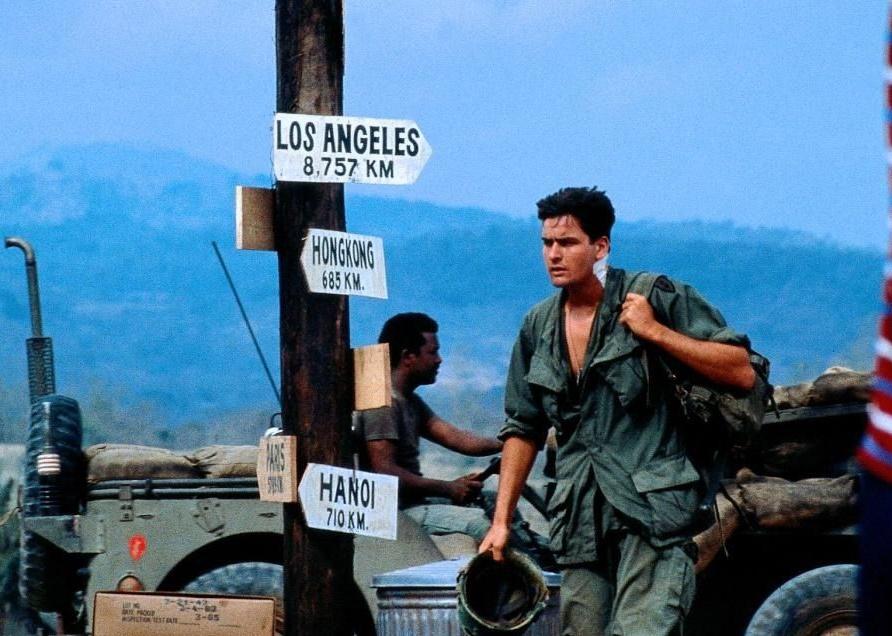 Charlie Sheen in military gear walking past a sign pointing to various cities with a military jeep in the background. 