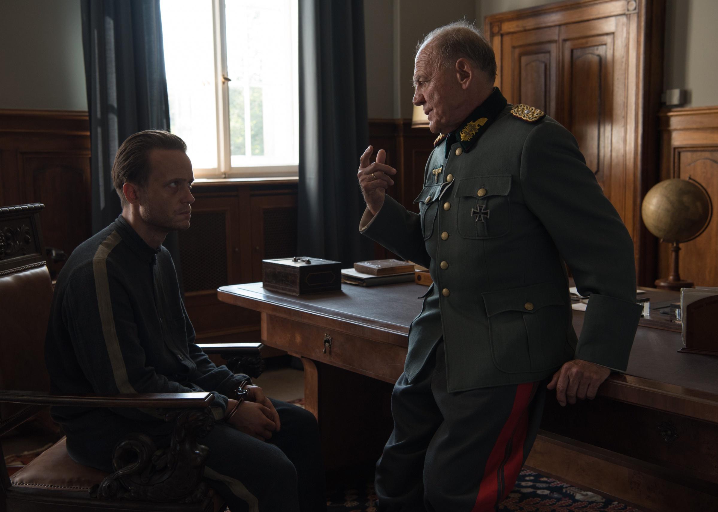 A military officer speaks to a man in handcuffs in a chair.