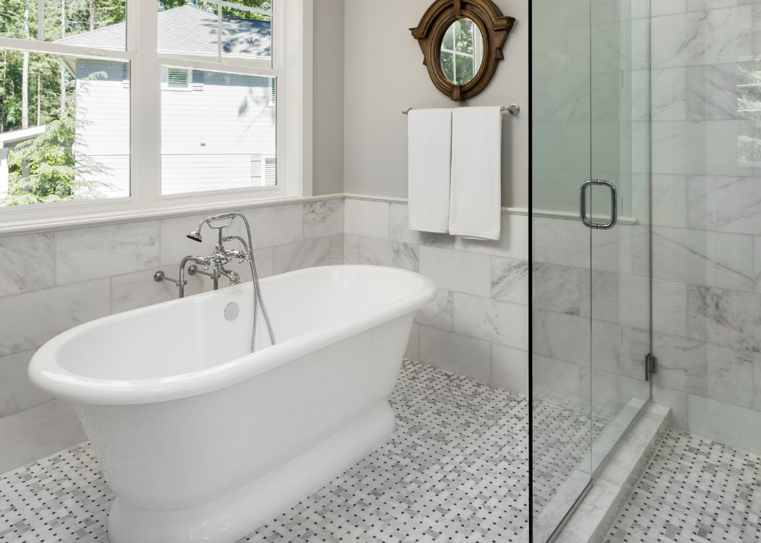 A bright grey and white bathroom with a white freestanding tub and glass enclosed shower. 