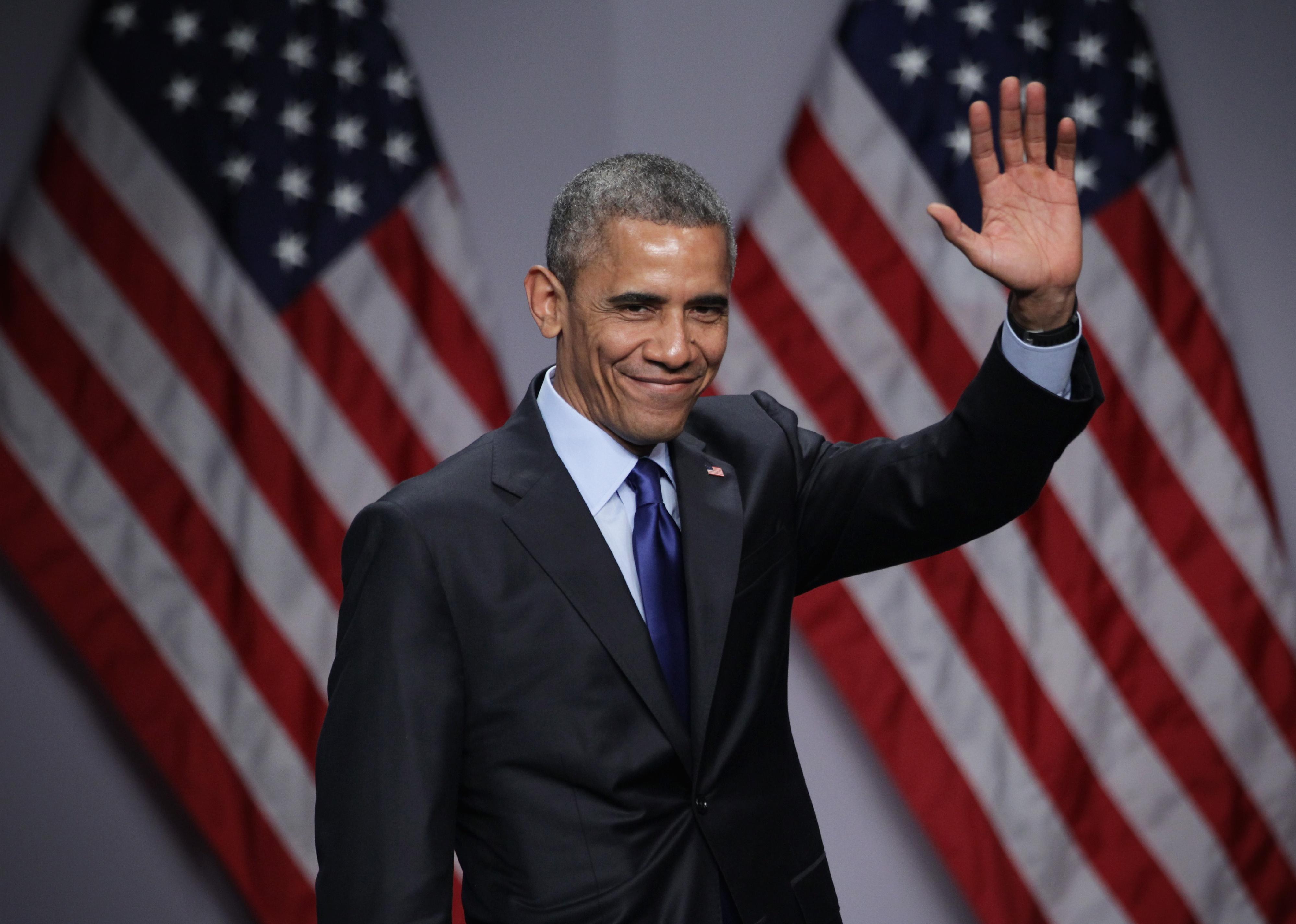 Barack Obama waving onstage in front of two American flags.