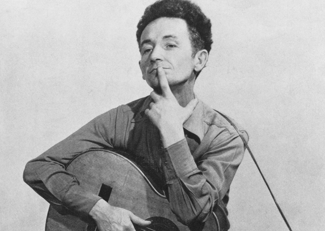 Woodie Guthrie holding a guitar with a finger over his mouth.