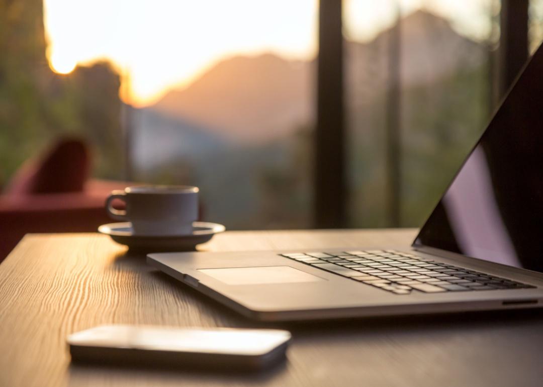 A laptop, a cup of coffee, and a phone on a table with a person blurred out in the background relaxing outside.