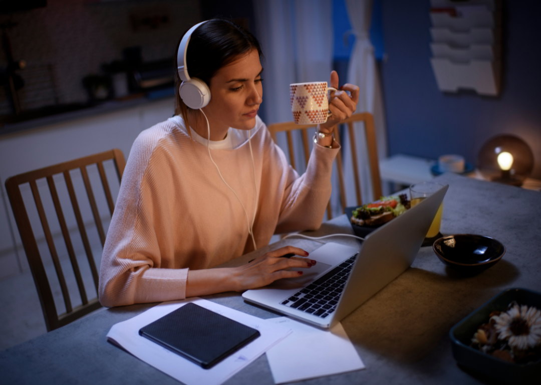 A woman working at her computer at night holding a cup of coffee and wearing headphones.