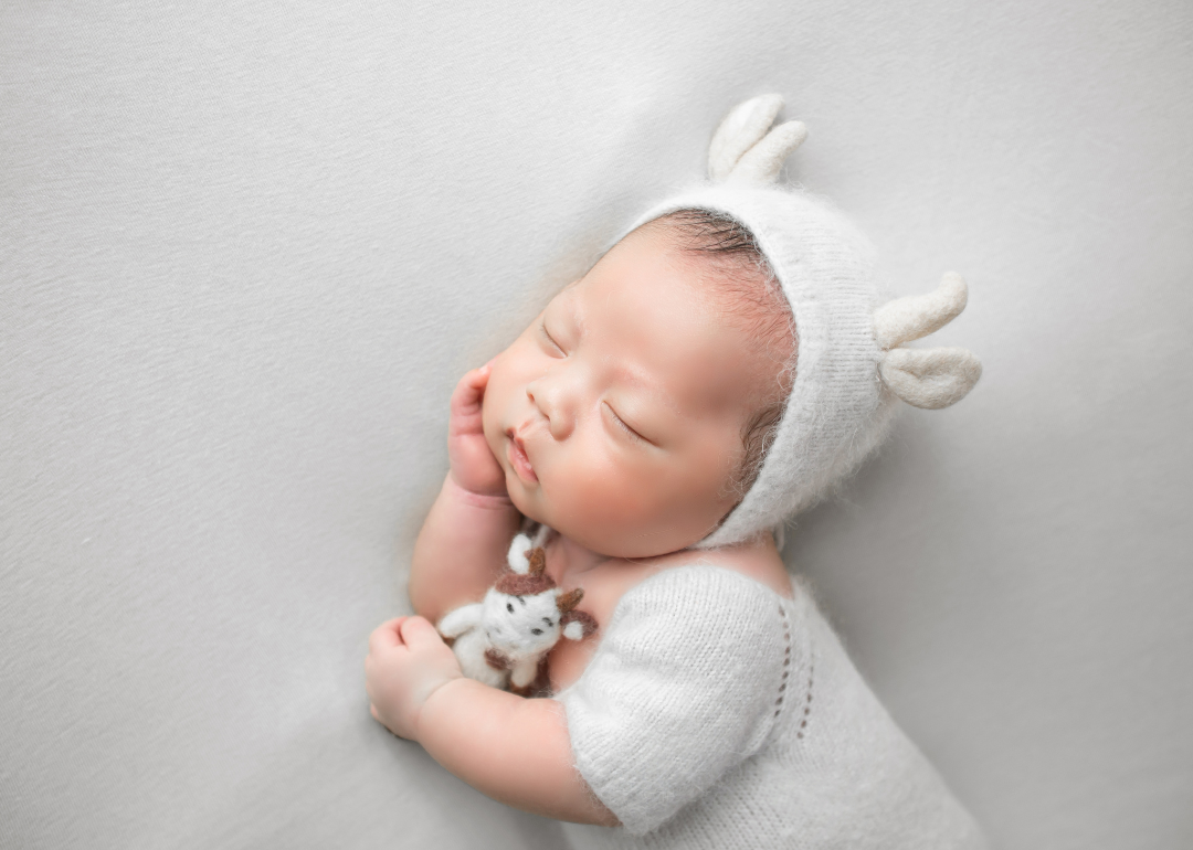 A sleeping baby wearing a fuzzy white outfit and hat with ears while cuddling a small brown and white cow.