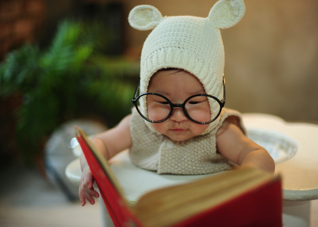 A baby wearing glasses and a hat with ears while holding a book open.