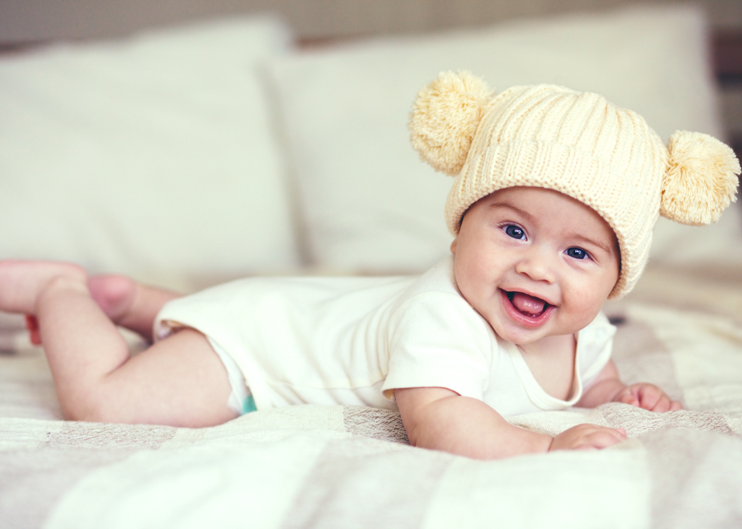 A baby smiling in a white onesie and hat with pom poms on the sides.