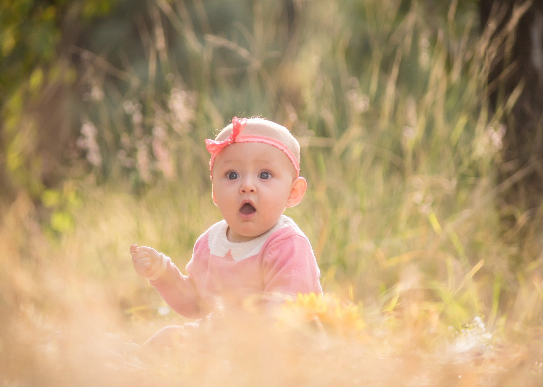 A baby with a surprised face sitting outside wearing a pink outfit and headband.