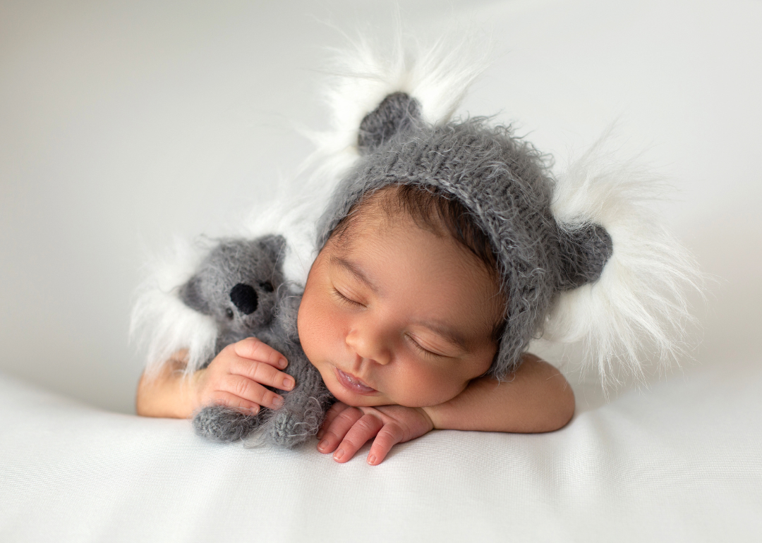 A sleeping baby in a gray and white furry hat holding a Koala bear.