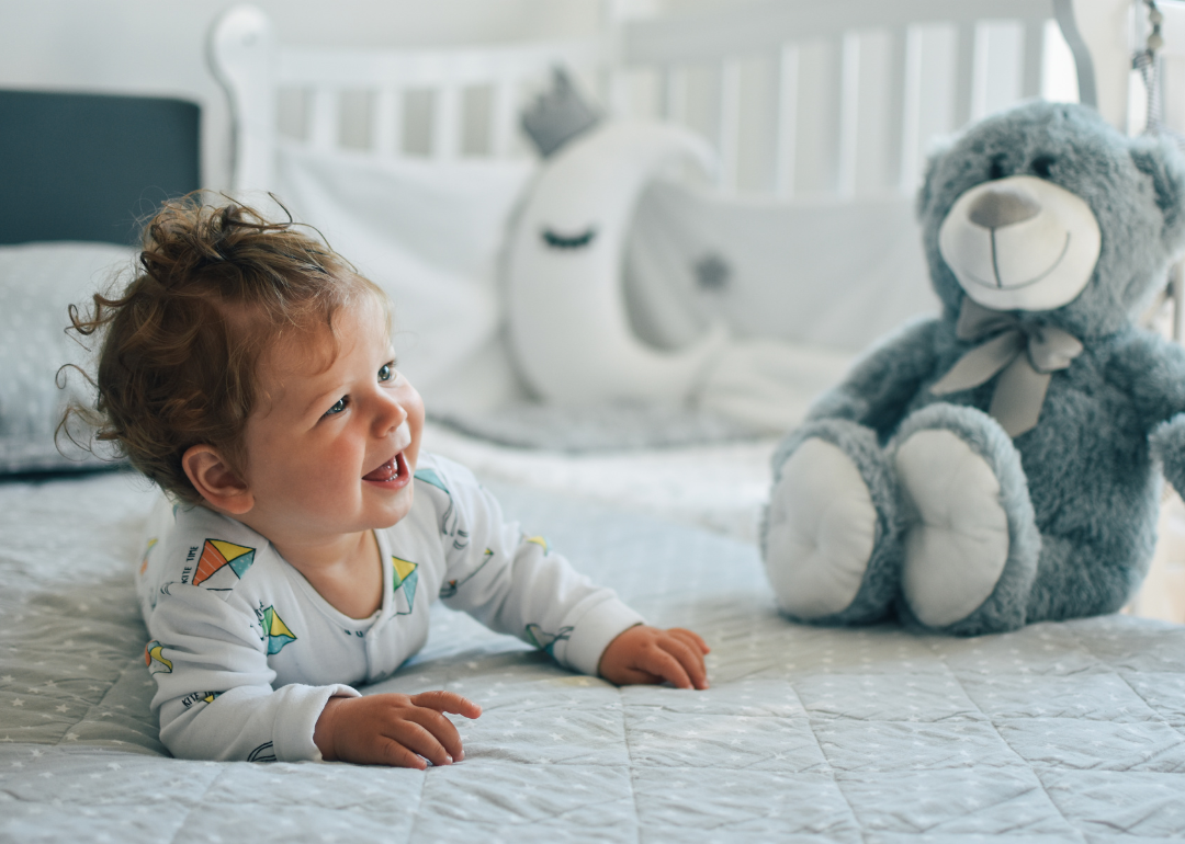 A baby in a kite onesie on a gray and white bed next to a gray bear.