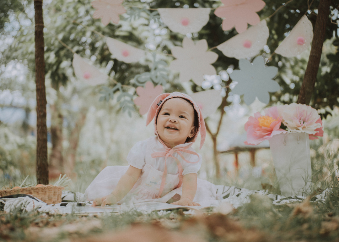 A baby posed on a blanket outside with paper flowers and paper flags hanging above.