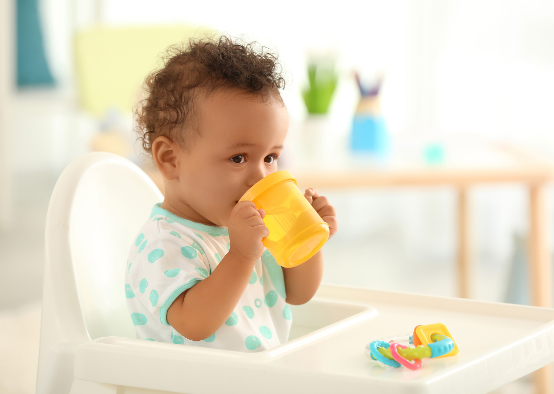 A baby in a high chair drinking from a yellow sippy cup.