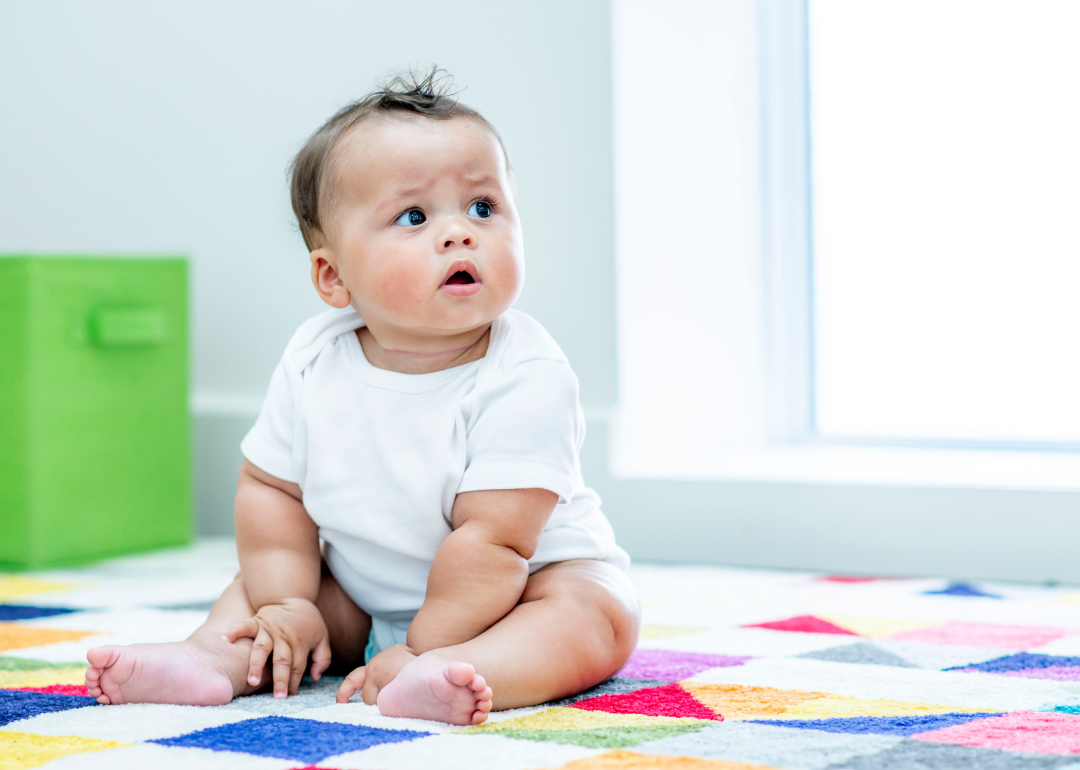 A baby boy in a white onesie on a blanket of colorful squares.