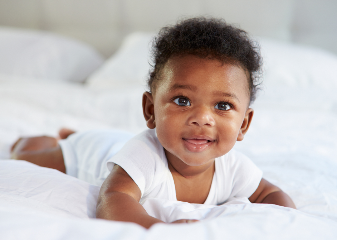 A smiling baby boy in a white onesie lying on a white fluffy blanket.