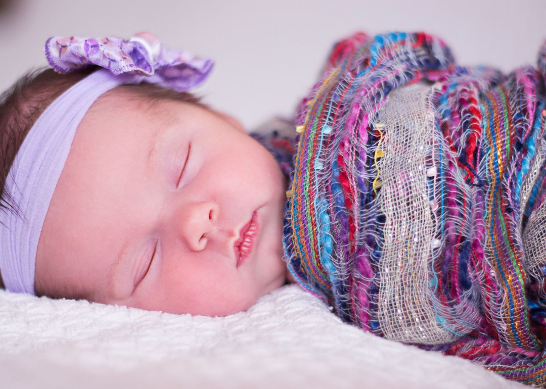 A sleeping baby girl wrapped in a colorful swaddle and wearing a purple headband.
