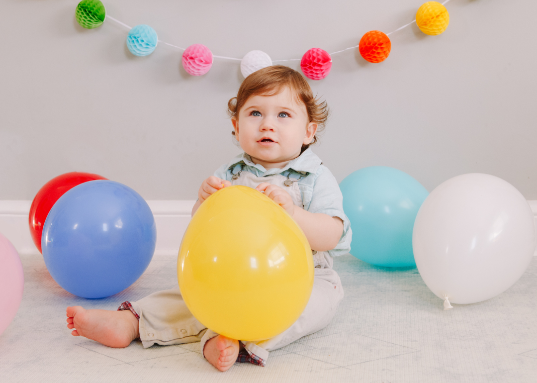 A baby boy in tan overalls sitting with colorful balloons.