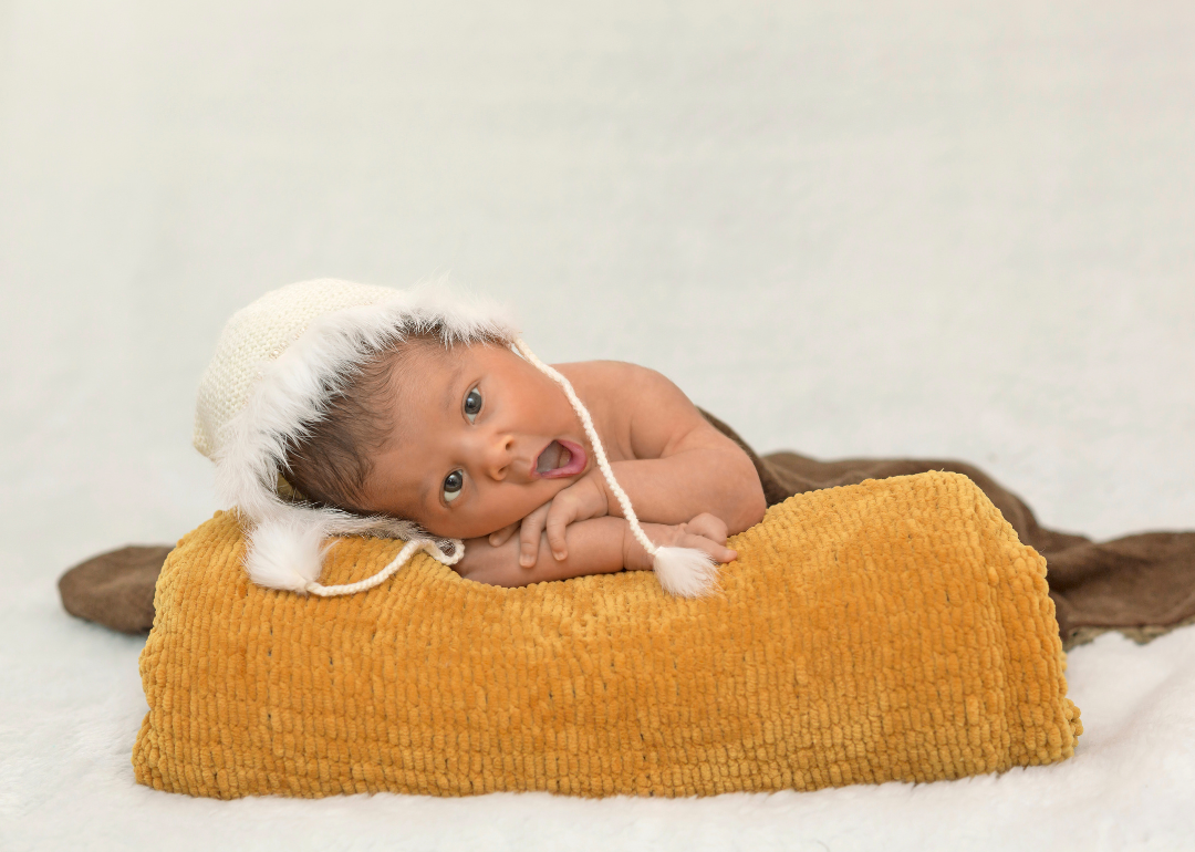 A baby wearing a hat and leaning over a fluffy pillow.