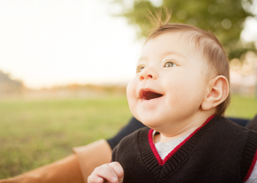 A smiling baby boy wearing a sweater vest outside.