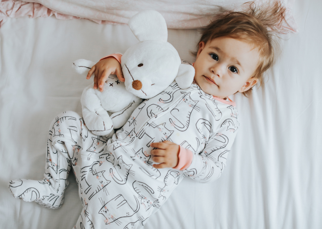 A baby girl in cat pajamas holding a stuffed animal.