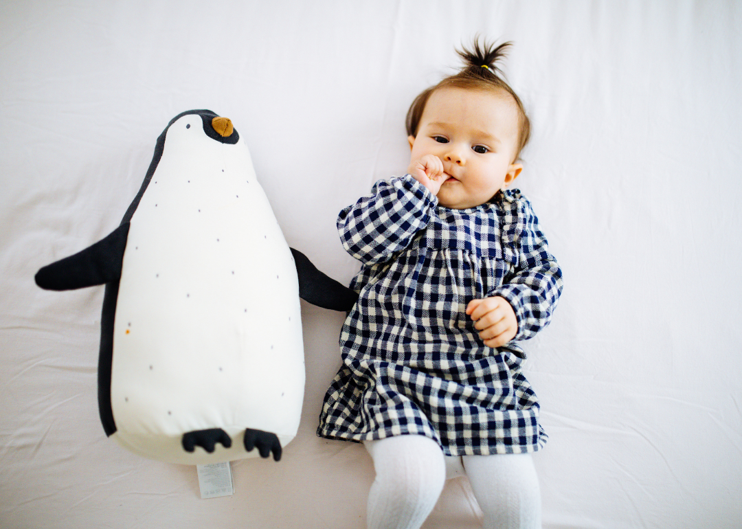 A baby girl in a blue and white checkered outfit lying next to a stuffed penguin.