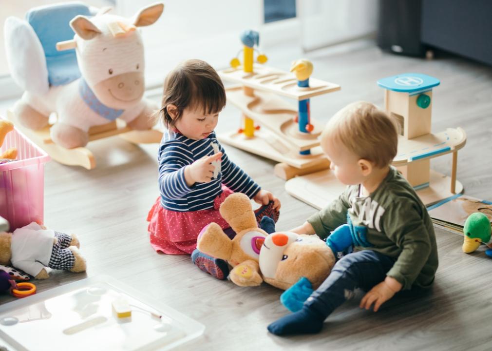 Two babies playing with a plush bear and surrounded by toys.
