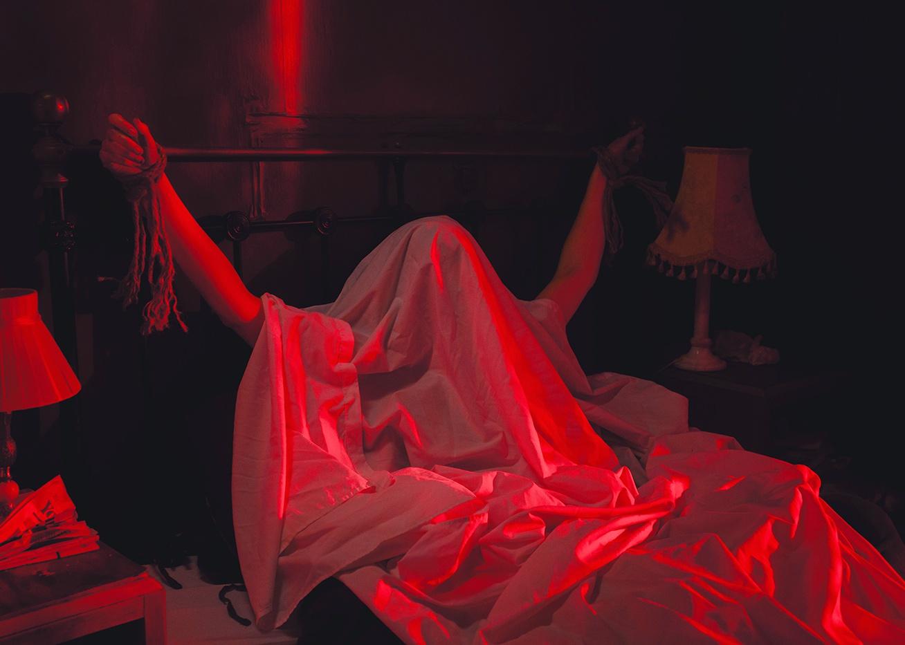 A person under a sheet tied to a bed in a red light