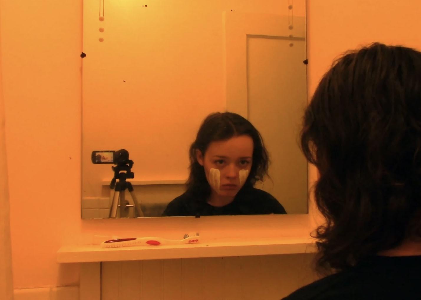 A young girl next to a tripod in her bathroom looking in the mirror with white paint down her cheeks