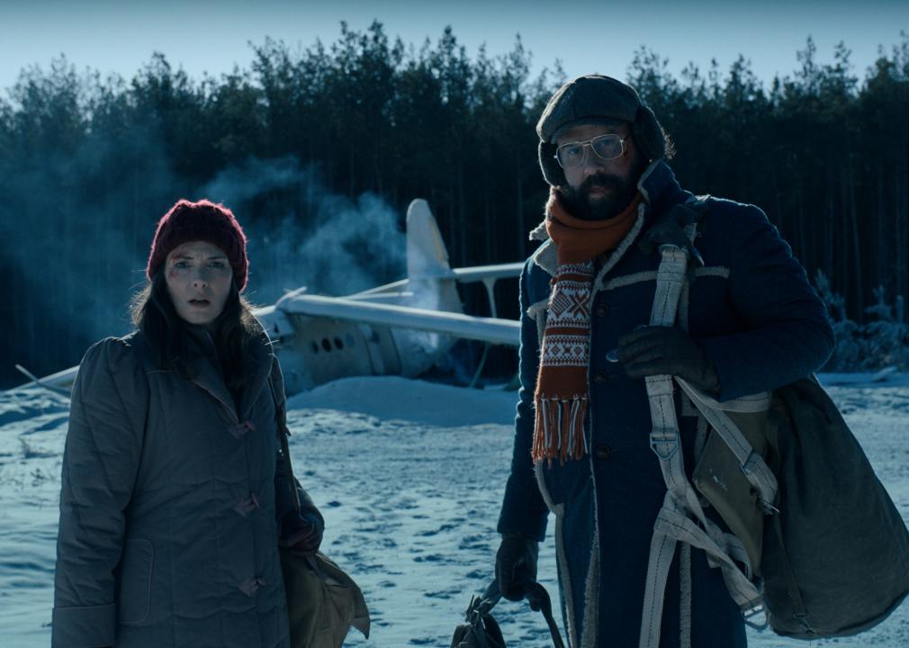 Winona Ryder and Brett Gelman standing in the snow in front of a plane crash.