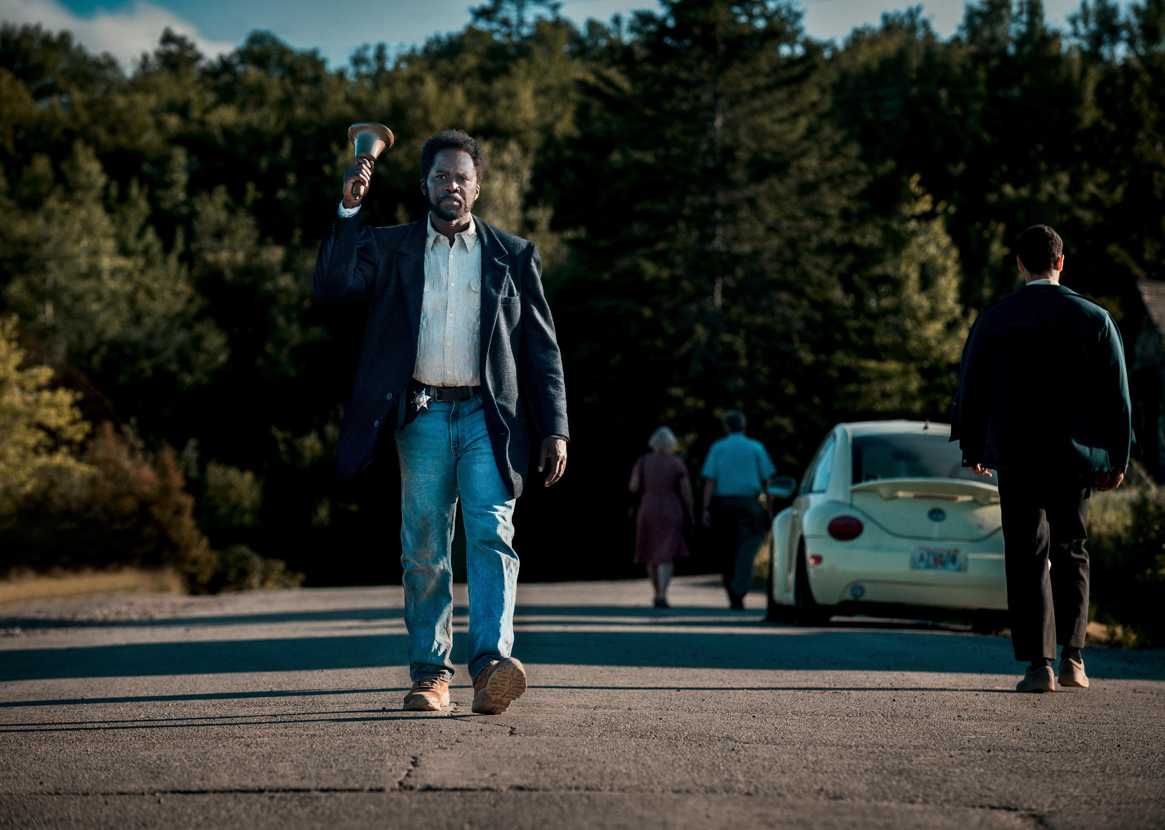 Harold Perrineau walking down a street ringing a bell and wearing a sheriff's badge.
