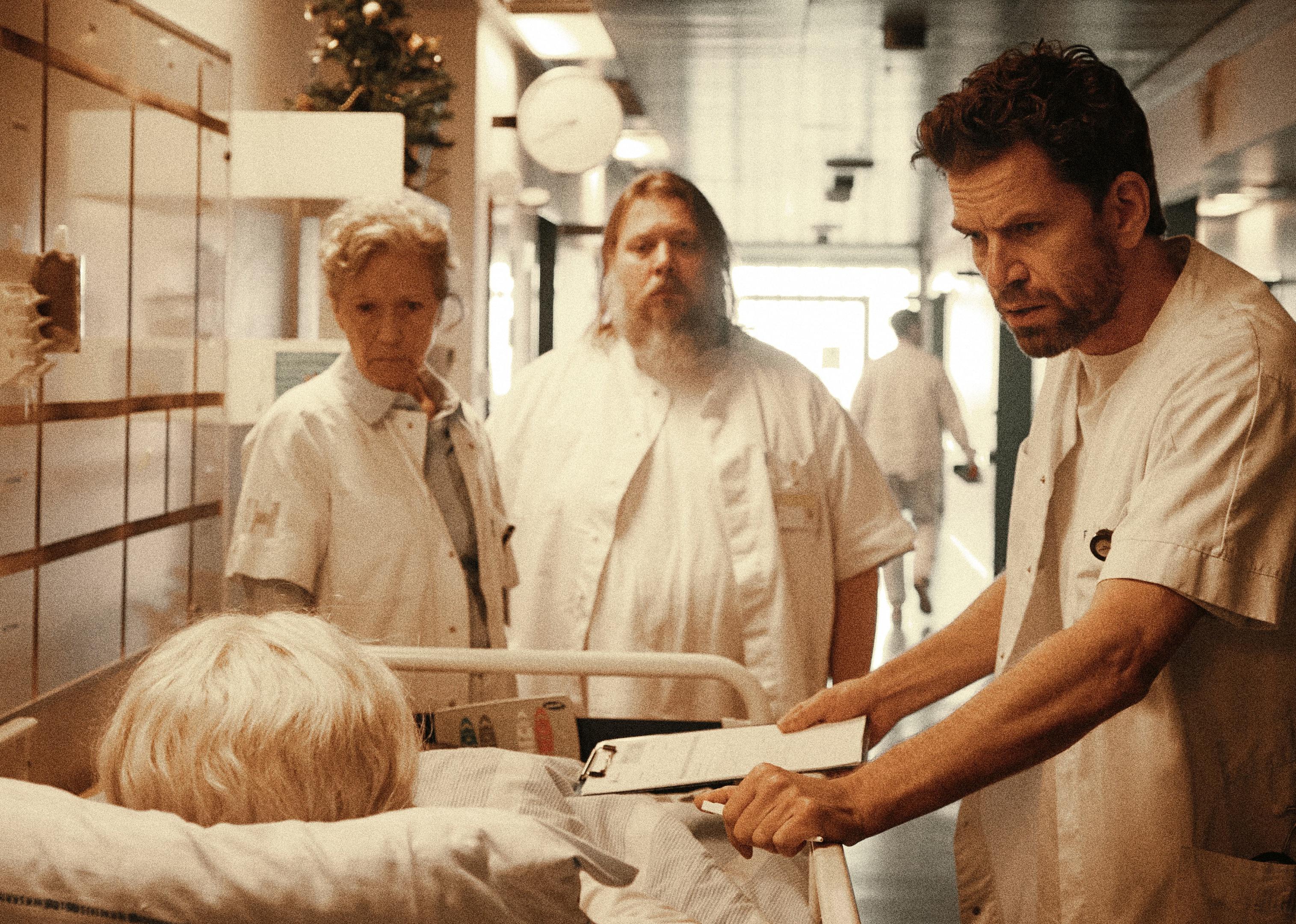 Three confused looking people talk to someone with white hair in a hospital bed in a hallway.