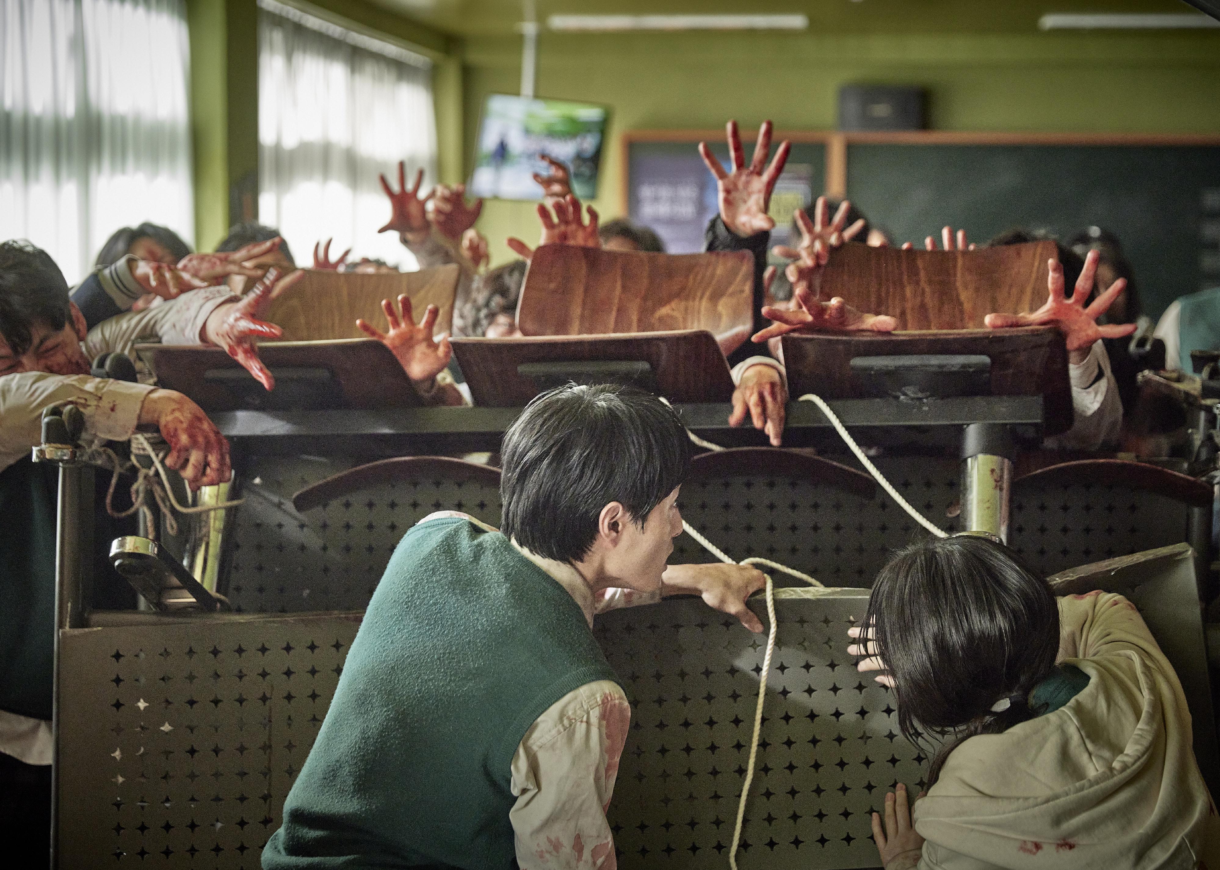 A man and woman holding down a barricade of desks in a classroom with hands trying to break over the top.