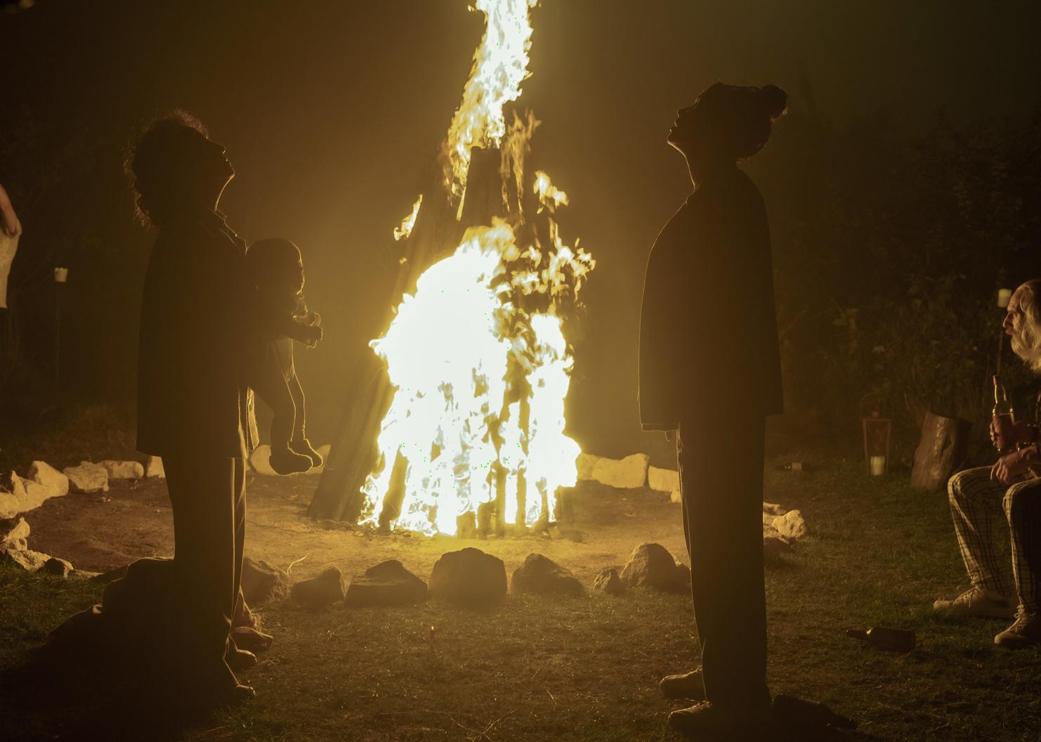 Two women standing next to a bonfire, one of them holding a baby as they look up.