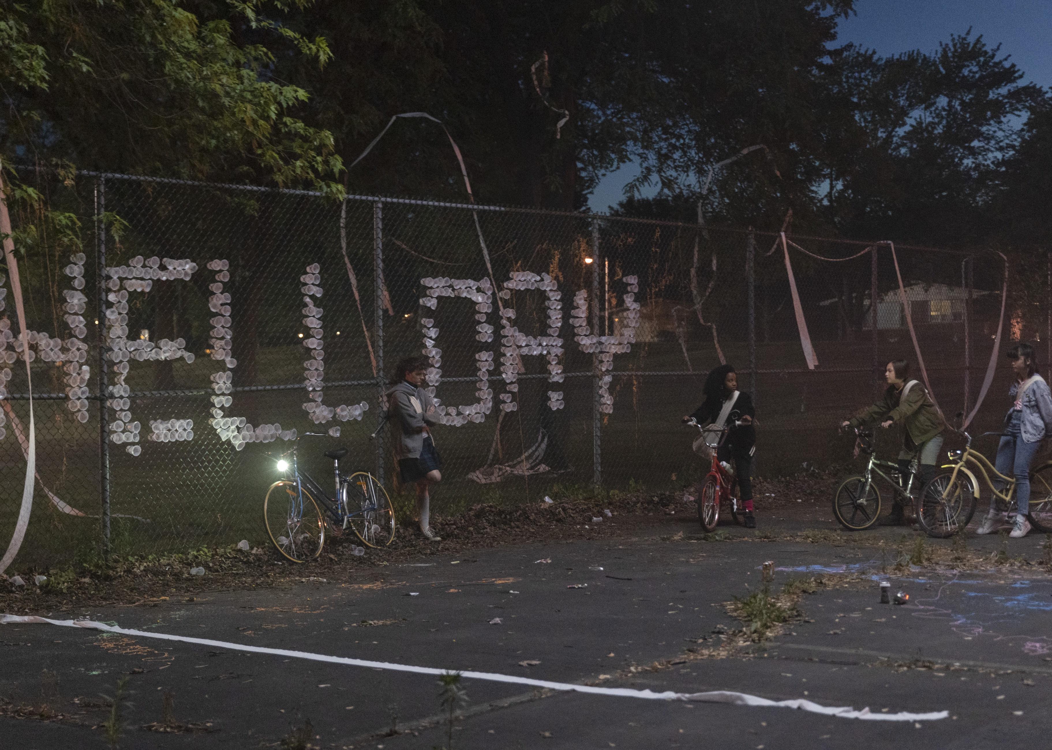 Four young disappointed girls with bikes next the message, "Hell Day", spelled out in cups on a fence.