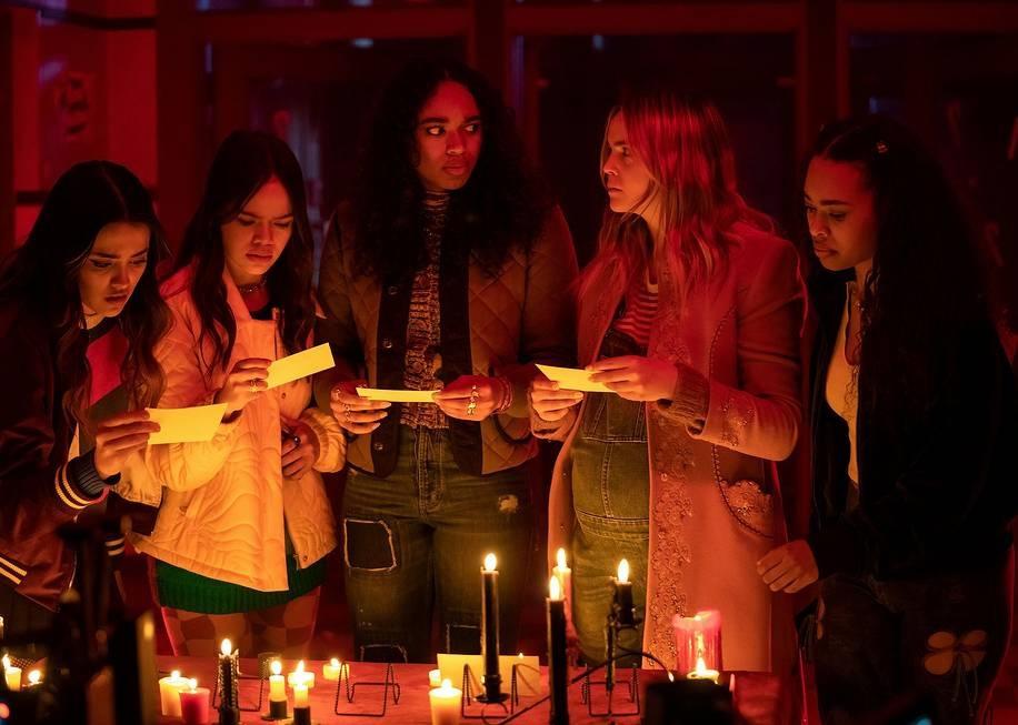 Five young women standing over a table of candles looking confusingly at cards.