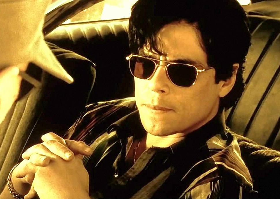 Benicio Del Toro wearing aviator sunglasses in a car talking to someone at his window.