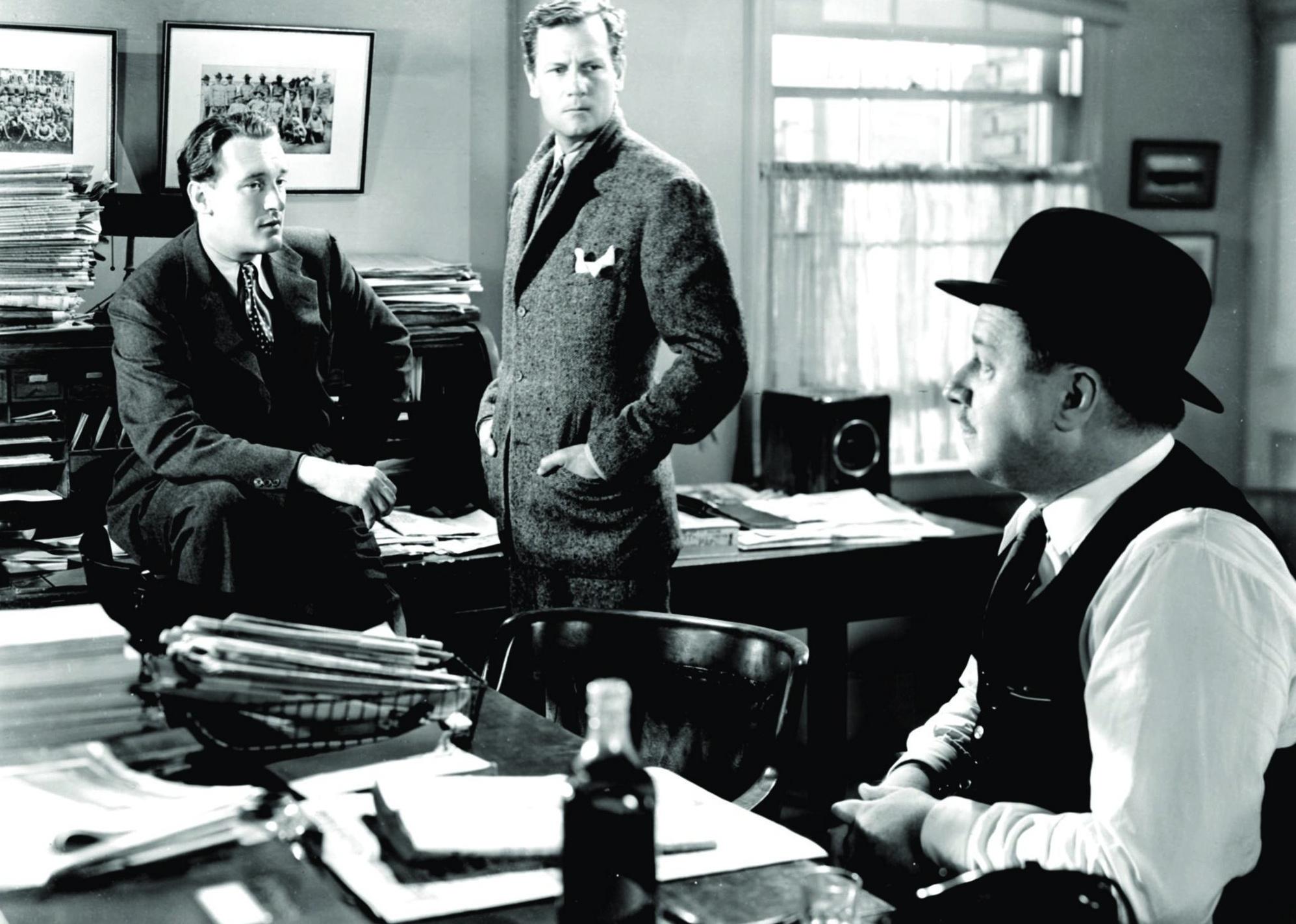 Two men in suits talk with a man sitting at a desk wearing a top hat.