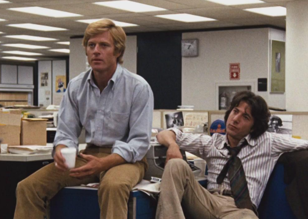 Dustin Hoffman and Robert Redford sit on top of desks in an office.